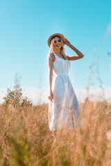 Fototapeta na wymiar selective focus of young blonde woman in white dress touching straw hat while posing in field against blue sky