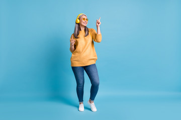 Full length photo of attractive hipster old woman listen music headphones direct fingers up energetic dancing alone wear sunglass yellow jumper jeans vivid blue color background