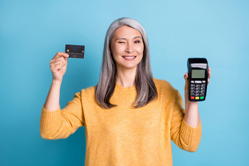 Photo of positive old woman hold credit card machine reader recommend bank purchase wink blink wear style stylish trendy sweater isolated over blue color background