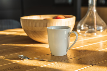 Cup of fresh coffee in a white cup and a wooden bowl with fruits on a wooden table in the kitchen in the morning sun