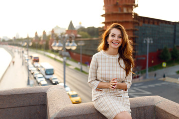 Cheerful elegant young woman with red hair wear e in styling beige dress standing on balcony, moscow city background.