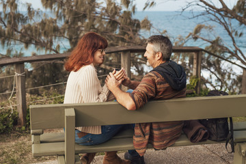 Happy middle-aged couple in love sitting on the bench in park, smile and drink tea or coffee from thermos.