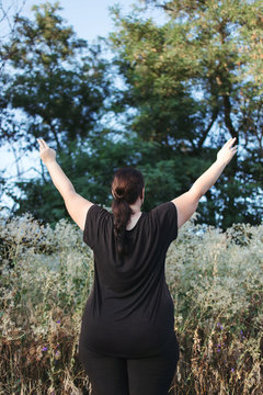 Body positive, freedom, high self esteem, confidence, happiness, obesity. Overweight woman rising hands towards the sky contemplating outdoors.