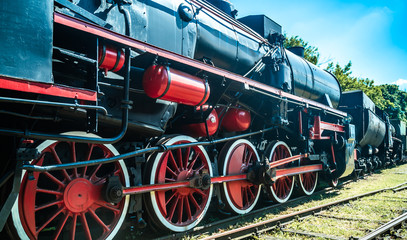 Naklejka premium Huge vintage steam locomotive, red painted steel wheel detail close up. Coal-powered steam train stands on a siding. Classic gigantic heavy railway machinery. Side view of power parts of machine.