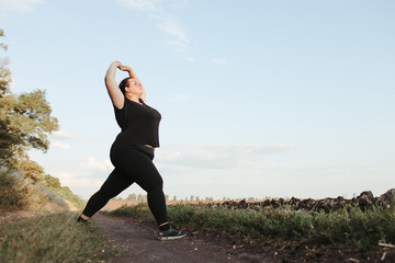 Young overweight woman doing morning yoga on the country road, open air activity. Healthy lifestyle, sport, weight loss
