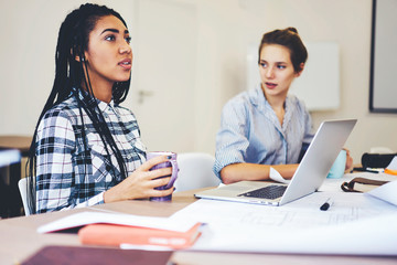 Young female pupil of business school answering on teacher question showing good knowledge in economics while her classmate creating report working on laptop computer connected to fast 5G wireless