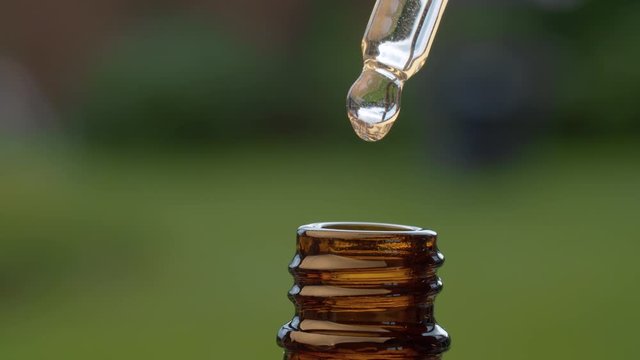 Macro shot essential oil dripping from a pipette on blurry natural background. Extreme close up hand taking transparent oil from a brown glass bottle