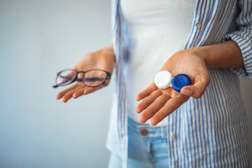 woman holding contact lens case on hand and holding in her other hand a glasses on grey background., eyesight and eyecare concept. Young woman choosing between contact lenses or glasses