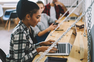 Young afro american copywriter in good mood chatting in social networks with followers and reading positive comments in own blog of design on laptop computer using 4G internet in coworking interior