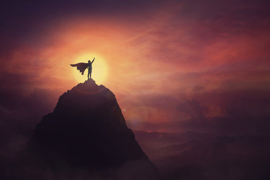 Conceptual sunset scene, superhero with cape standing brave on top of a mountain looks determined at horizon raising one hand up as a winning leader. Hero power and motivation, overcoming obstacles.