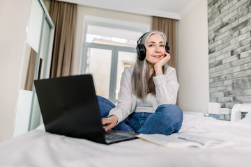 Charming elderly gray haired woman in headphones, wearing jeans and white shirt, typing on the laptop, having fun with friends or working online, while sitting on the bed