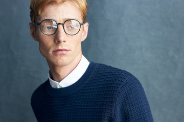 portrait of a young handsome red-haired man with freckles on a dark background he is wearing a classic dark blue sweater on a white shirt and has transparent glasses with a black frame