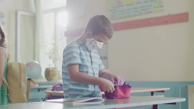 A pupil of elementary school packing up a stuff and wearing a protective mask. Post quarantine school, new normality, new rules, social distance concept.