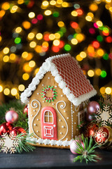 Gingerbread house with pine branches and chrismtas decorations on dark wooden table, chrismtas lights in background, selective focus