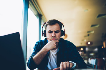 Young handsome pondering male student dressed in trendy clothing enjoying recreation with modern technology sitting in coffee shop.Pensive hipster guy spending leisure time listening to online course