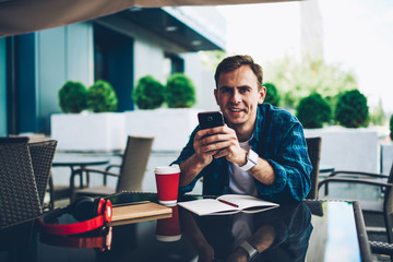 Half length portrait of handsome smiling male student chatting with followers in social networks during free time outside.Young happy man looking at camera while enjoying recreation with device