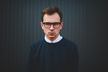 Obraz premium Half length portrait of handsome confident male person in stylish eyeglasses spending free time at urban setting.Young hipster guy looking at camera during posing against wall at summer street