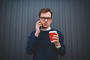 Portrait of young male person in cool spectacles having mobile conversation via smartphone during walk next to promotional background.Confident hipster guy looking at camera while talking on phone