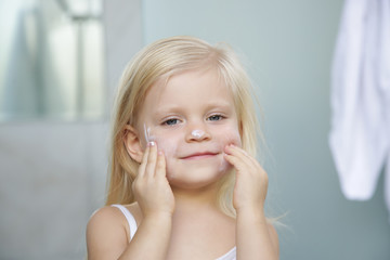 Adorable blonde caucasian toddler girl applying cream on her face	