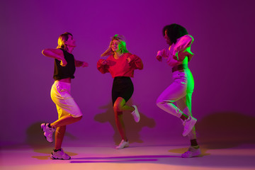 Flexible. Sportive girls dancing hip-hop in stylish clothes on purple-pink background at dance hall in green neon light. Youth culture, movement, style and fashion, action. Fashionable portrait.
