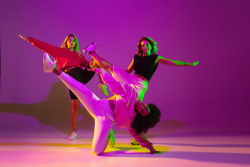 Flexible. Sportive girls dancing hip-hop in stylish clothes on purple-pink background at dance hall in green neon light. Youth culture, movement, style and fashion, action. Fashionable portrait.