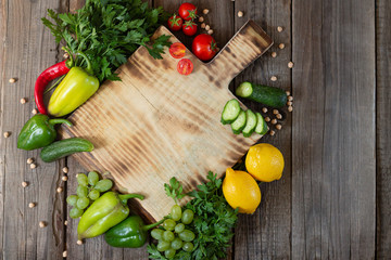 Wooden cutting board next to fresh herbs, raw vgetables and fruits on rustic wood table top view