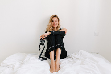 Naklejka premium Full-lenght photo of lovely tender girl with blonde hair wearing home suit sitting on the white bed with smile and posing at camera. Happy morning at home