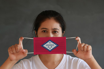A woman and hygienic mask with Arkansas flag pattern in her hand and raises it to cover her face. A...