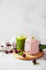 Fresh herb smoothies and cherry smoothies in glass jars sit on a wooden stand against a concrete backdrop.