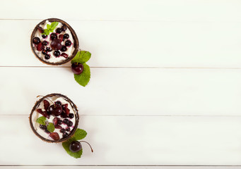 Dessert of whipped cream and fresh berries of currant and sweet cherry with mint leaves in coconut cups on a white wooden background. Flatlay. Copy spaes.