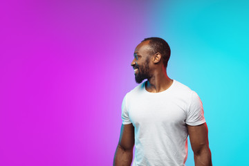 Smiling. African-american young man's portrait on gradient studio background in neon. Beautiful male model in casual style, white shirt. Concept of human emotions, facial expression, sales, ad.