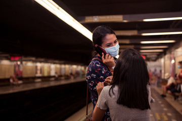 Young woman and her daughter on the subway station. Woman wearing face mask and talking on the smartphone while her daughter claim attention.