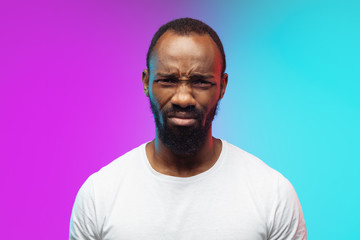 Close up offended. African-american young man's portrait on gradient background in neon. Beautiful male model in casual style, white shirt. Concept of human emotions, facial expression, sales, ad.