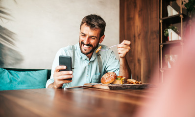 Handsome middle age man sitting in restaurant and enjoying in delicious burger. He is happy and smiled and uses his smart phone to talk with someone.