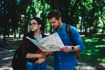 Male traveller with backpack asking attractive student in eyeglasses and book in hands about right direction of location on map standing outdoors on street.Couple of tourist searching rout during trip