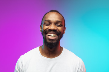 Close up laughting. African-american young man's portrait on gradient background in neon. Beautiful male model in casual style, white shirt. Concept of human emotions, facial expression, sales, ad.