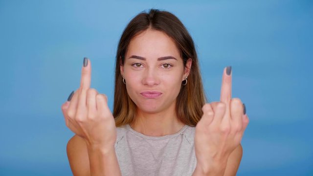 Positive woman in gray t-shirt smiles, dances, shows middle finger on blue background with copy space. 4K slow motion footage