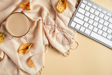 Autumn flat lay composition with fallen leaves, scarf, coffee cup, computer keyboard, glasses on beige background. Cozy home office desk, feminine workspace. Hygge style.