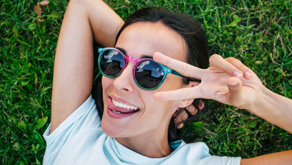 Happy beautiful hipster woman in cool sunglasses is having fun while she is lying on the lawn and looking on camera