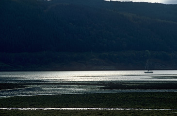 Loch Sunart in Scottish Highlands UK. Late afternoon. Evening sunset reflected on water surface. Single sailing boat. Mountains and forests in background. Tidal streams.