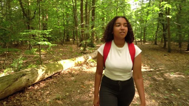 Teenage mixed race African American girl young woman hiking with a red backpack and using a cell phone in forest woodland 