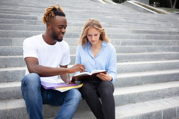 Education, learning and people concept - young two multiracial friends sitting on stairs in the park, study for their exams and look at the lecture notes.