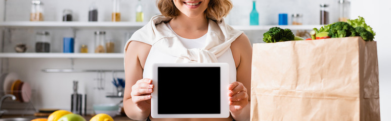 panoramic crop of woman holding digital tablet with blank screen near paper bag with groceries