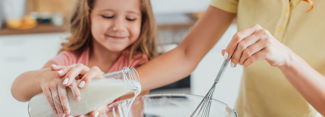 cropped view of mother holding whisk and pouring milk into glass bowl together with daughter,...