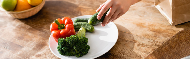 panoramic orientation of young woman holding cucumber near plate with broccoli and bell pepper