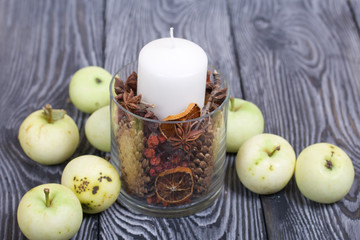 A candle in a glass candlestick. The candlestick is filled with dried cones, spices and dried fruits. Apples lie on the table surface nearby.