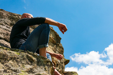 A young violent man is sitting on the edge of a cliff in the mountains. A strong athlete with a shaved head sits on a rock view from below against the sky.