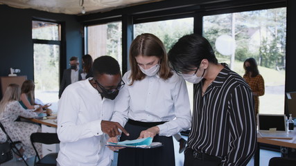 Young happy diverse gen-z business colleagues work together, all wearing COVID-19 face masks at modern light office.