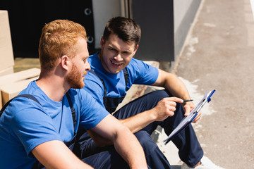 Obraz premium Loaders with pen and clipboard talking while sitting in warehouse outdoors