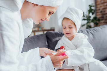 selective focus of young woman making manicure to daughter while sitting together in white bathrobes and towels on heads